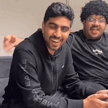 two men are sitting on a couch and one has an eagle sweatshirt on