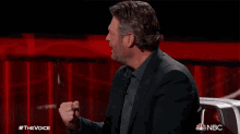 a man in a suit is sitting in front of a red curtain with the words #thevoice on the bottom