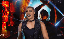a woman in a black vest with spikes on it is standing on a stage in front of a sign that says nxt