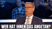 a man in a suit and tie is sitting in front of a crowd and says " wer hat ihnen das angetan " .