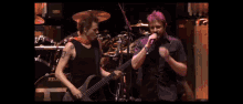 a man playing a guitar next to another man singing on a stage with a tama drum in the background