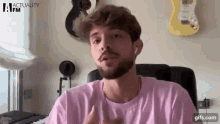a man with a beard wearing a pink shirt is sitting in front of a microphone in a room .