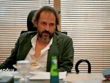 a man with a beard is sitting at a desk in front of a window with blinds and the word moglu on the bottom