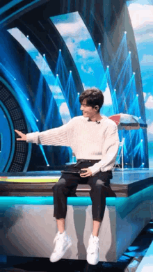 a man in a white sweater is sitting on a stage in front of a blue background