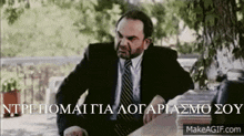 a man in a suit and tie is sitting at a table with the words makeagif.com above him .
