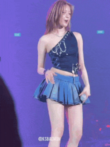 a woman in a black top and blue pleated skirt is holding a microphone and has the hashtag @ksbaemon on the bottom