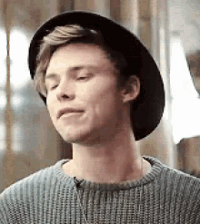 a young man wearing a hat and a sweater is making a funny face with his eyes closed .