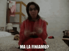 a woman in a red jacket is sitting on a bed with the words ma la finiamo written on the bottom of her face .