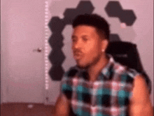 a man in a plaid shirt is sitting in a chair making a funny face .