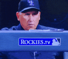 a man wearing a rockies hat sits in front of a rockies.tv sign