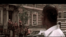 a man in a white shirt is standing in front of a building with a group of people standing on the sidewalk .