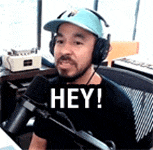 a man wearing headphones and a hat is sitting in front of a microphone and says hey .