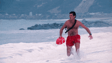 a shirtless man in red shorts is running in the water holding a red life preserver