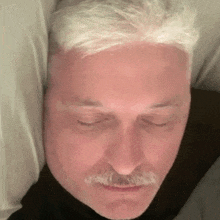 a man with white hair and a mustache is laying in bed with his eyes closed .