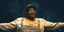 a man wearing overalls and a cowboy hat is smiling with his arms outstretched