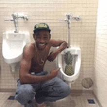 a shirtless man is squatting in front of a urinal with a pineapple in it .