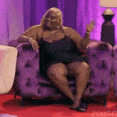 a woman in a black dress is sitting in a purple chair and waving .