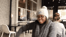 a woman wearing a white hat and glasses is sitting at a table outside a restaurant .