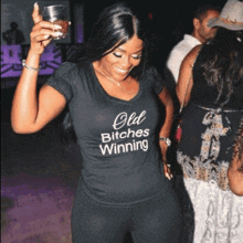 a woman wearing an old bitches winning shirt holds a glass