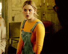 a woman wearing overalls and an orange shirt is standing in a dark room