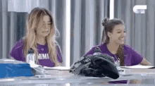two women in purple shirts are sitting at a table with a bottle of water .