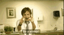 a man in a white shirt and tie is standing in a kitchen with the words `` be a winner '' on the screen .