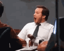 a man in a suit and tie is sitting at a table with his mouth open and a surprised look on his face .