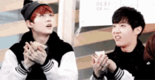 two young men are sitting next to each other eating sandwiches and clapping .