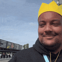 a man wearing a yellow crown smiles in front of a billboard that says ' a ' on it