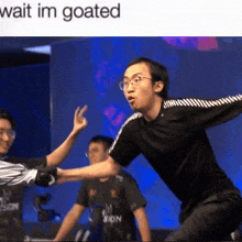 a man in a black shirt with a white stripe on the sleeves stands in front of a sign that says wait im goated