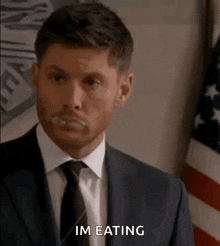 a man in a suit and tie is eating something in front of an american flag .