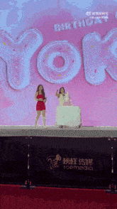 two women standing on a stage with a cake in front of a sign that says " birthday "