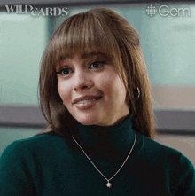 a woman wearing a green turtleneck and a gold necklace with a gem logo in the background