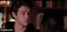 a young man is standing in front of a bookshelf and talking to a woman .
