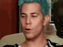 a man with blue hair and a tattoo on his forehead is sitting on a couch .