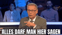 a man in a suit and tie is pointing at something in front of a crowd of people and says alles darf man hier sagen .