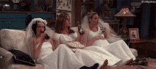 three women in wedding dresses are sitting on a couch drinking wine and eating popcorn .