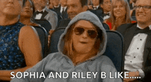 a woman wearing a hoodie and sunglasses is sitting in a crowd of people at an event .