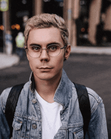a man wearing glasses and a denim jacket has a backpack on his back