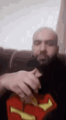 a bald man with a beard is sitting on a couch wearing a superman shirt and smoking a cigarette .