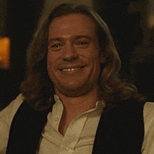 a man with long hair is laughing and wearing a vest