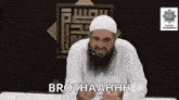 a man with a beard and a white hat is sitting at a table with a microphone .