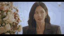 a woman in a black suit is standing in front of a vase of flowers and making a funny face .
