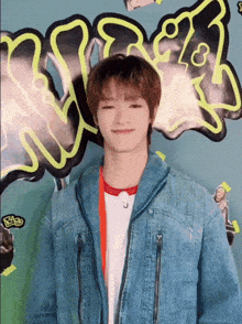 a young man wearing a denim jacket stands in front of a graffiti wall