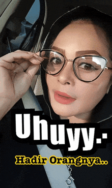 a woman wearing glasses is sitting in a car with the words uhuyy .. hadir orangnya
