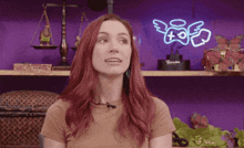 a woman with red hair stands in front of a purple wall with a neon sign that says ' angels ' on it