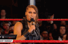 a woman stands in a wrestling ring holding a microphone and a sign that says ' very thursday '
