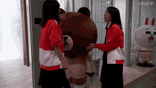 two women hugging a brown teddy bear in front of a sign that says fever on it