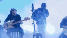 a man playing a guitar in front of a man wearing a jacket that says " xlarge "