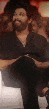 a man with a beard is sitting on a couch with his legs crossed and a necklace around his neck .